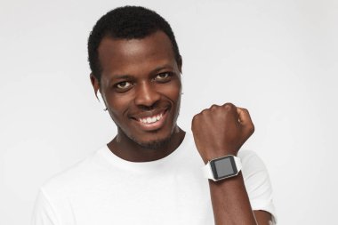 Afro-Amerikan yakışıklı beyaz casual T-shirt giyen ve bilek emin gülümseme ile akıllı kol saati gösteren gri arka plan üzerinde izole kapalı portre onun yaşam tarzı ile memnun