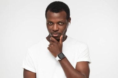 Gri arka planı izole yatay closeup genç yakışıklı Afrika adam resimde boş T-shirt ve giyen elektronik akıllı izlemek çene sanki bir şey tahmin etmek tırmalamak giymiş