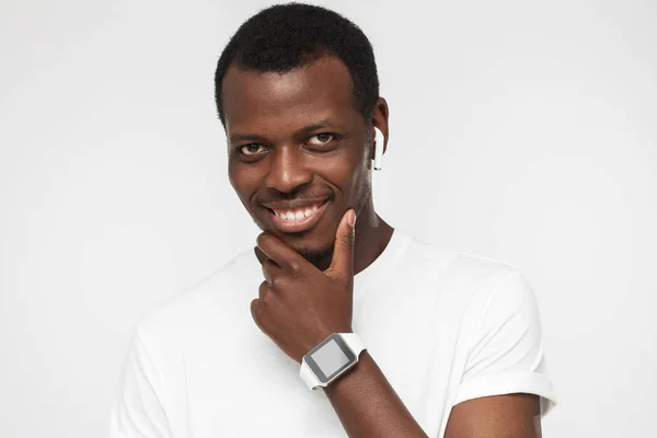 Indoor picture of young dark-skinned man isolated on gray background looking straight at camera, trendy wristwatch visible on wrist, showing positive smile to viewer feeling confident and relaxed