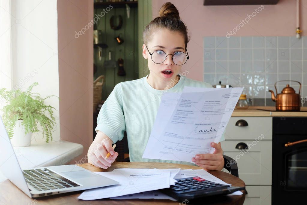 Horizontal closeup of European Caucasian lady spending afternoon at kitchen table, wearing eyeglasses, looking shocked with what she sees in documents she is dealing with, dissatisfied with debts