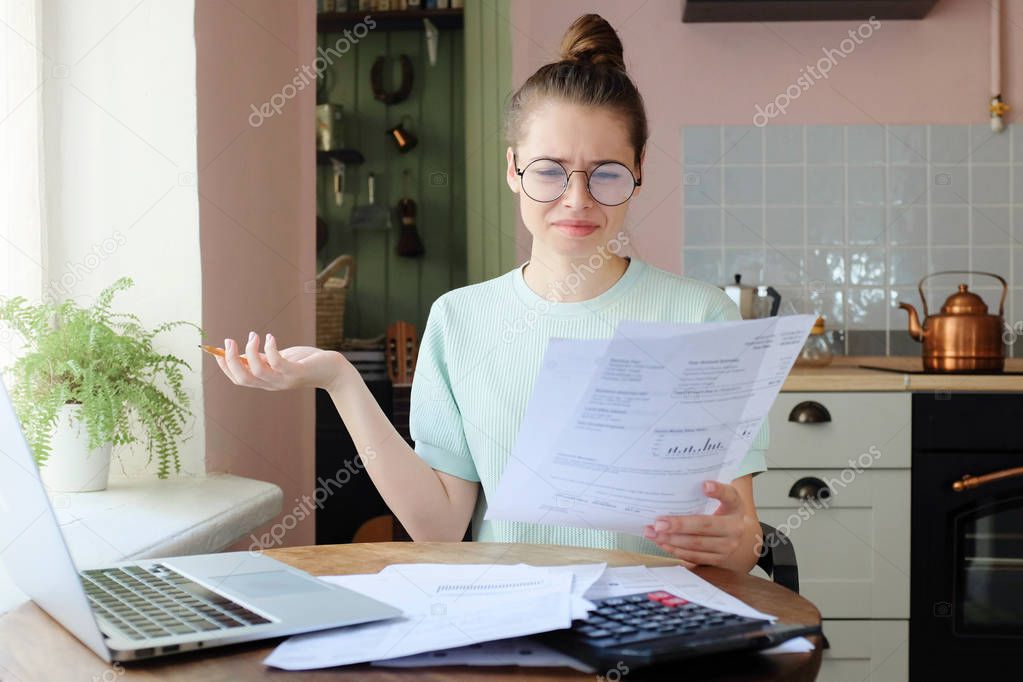 Closeup photo of young good-looking woman dressed in casual clothes staying in kitchen at home studying received bills with puzzled and helpless look and corresponding gesture with upset face