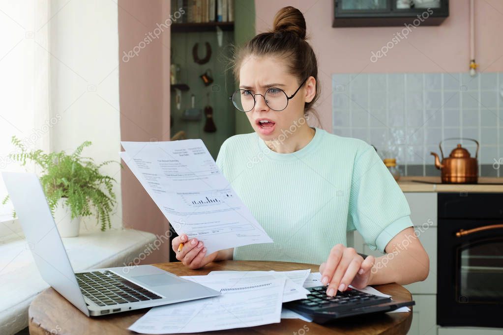 Indoor closeup of young emotional European lady dealing with sheets of paper and bills at home having opened mouth as if she can not believe these big numbers to pay are true, feeling despair