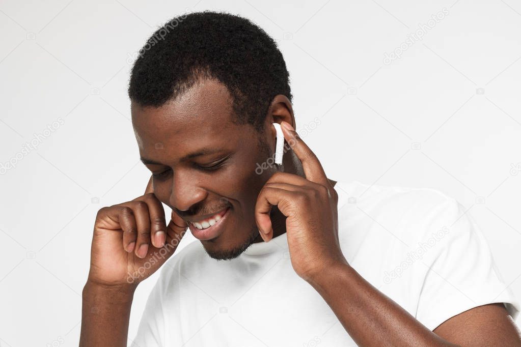 Closeup picture of African American man pictured isolated on gray background listening to player with happy toothy smile pressing hands to earphones to enjoy sound more, moving, feeling great joy