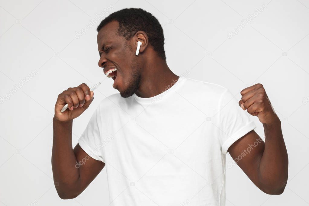 Horizontal photo of young African American man pictured isolated on gray background enjoying music through wireless earphones so much that he is dancing with closed eyes, energetic and full of joy