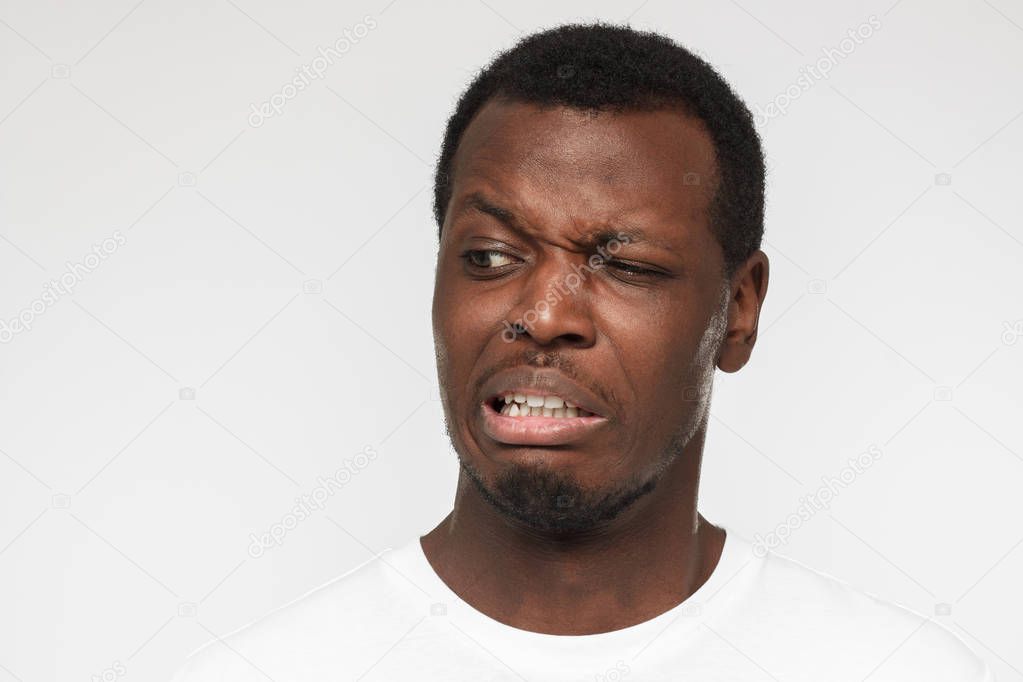 Studio photo of African American man dressed in casual T-shirt standing against grey background frowning and grinning with expression of strong disgust, absolutely dissatisfied with what he perceives