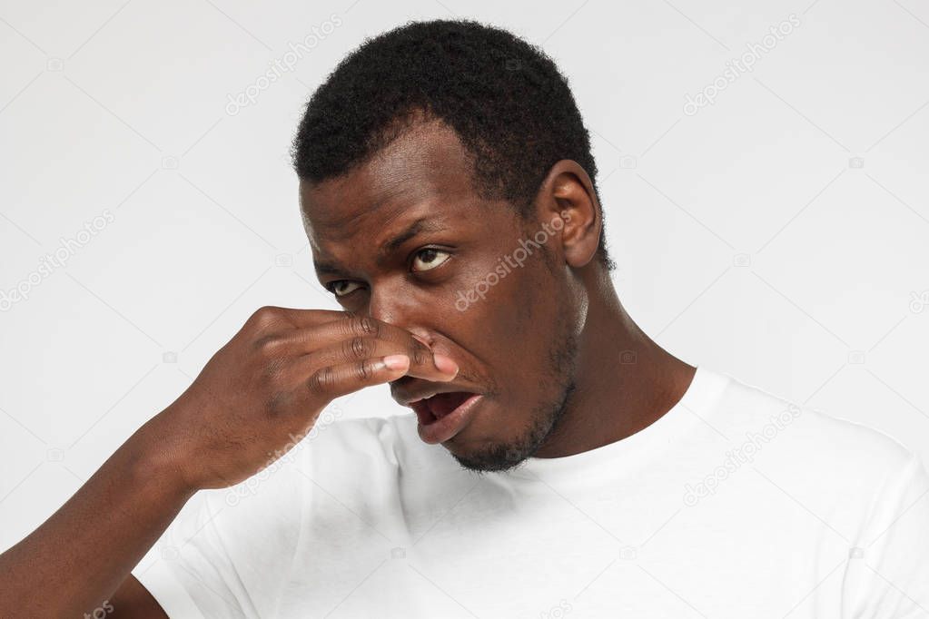 Horizontal indoor headshot of young African American man pictured isolated on gray background holding his nose because of unpleasant stinky odor he feels showing vivid face expression of disgust
