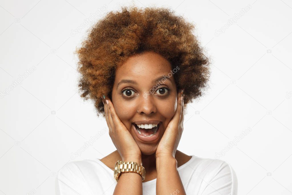 Young beautiful african american female isolated on gray background screaming happily in great astonishment, her eyes and mouth open eyed, palms pressed to her cheeks