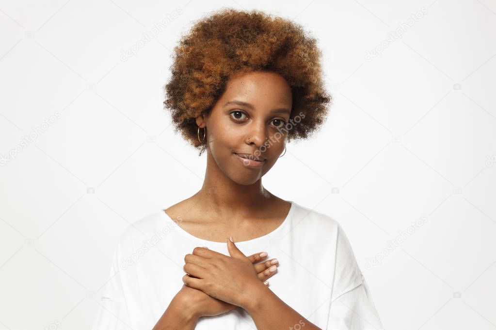 Portrait of smiling young woman keeps hands on chest, expresses sympathy. Kind hearted friendly african american female shows kindness, wearing white t-shirt