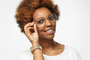 Beyaz t gömlek giymiş ve altın gözlüklerini gri arka plan üzerinde izole bir eliyle tutan genç gülümseyen neşeli koyu tenli Afrika kökenli Amerikalı kadın portresi stüdyo