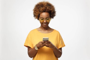 Sarı tshirt ve modaya uygun gözlük cep telefonu ekranına bakarak, web sayfalarını tarama ve güzel sohbet ederken gülümseyen gri arka plan üzerinde izole genç Afro-Amerikan kadın 