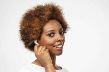 Onlardan daha iyi dinleme deneyimi sağlamak için dokunaklı beyaz kablosuz kulaklık giyen gülümseyen, kamera, yarı-açık gri arka plan üzerinde izole güzel esmer Bayan portre resmi