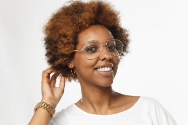 Dikkat et, kulağına küpe ve saçlarını dokunmaktan ve geniş gülümseme ile doğrudan kameraya bakarak burun gözlük, giyen gri arka plan üzerinde izole genç Afro-Amerikan Kız yatay resim