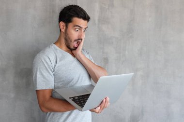 Dizüstü bilgisayar ekranına ağız ile seyir yakışıklı genç tur derin şaşkınlık ve şok, dokulu gri duvara karşı ayakta nedeniyle açık