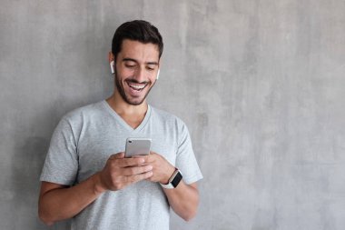 Yakışıklı adam gülerek mutlu bir şekilde her iki elinde tutuyor Smartphone ekranında içeriğe göre eğlendirdi gri rahat t-shirt giyen kapalı yaz resmi dokulu duvar karşı ayakta