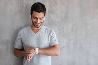 Kopya alanı ile gri dokulu duvar karşı ayakta dokunmatik ekranlı akıllı kontrol kablosuz kulaklık ve t tişört giyen genç yakışıklı adam izliyor 
