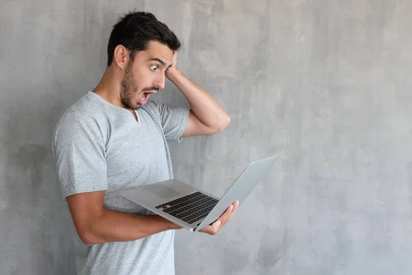 英俊的男子在灰色 T恤看着笔记本电脑屏幕 惊讶于大的销售价格 握手在头上 站在墙上与复制空间的质感墙壁 — 图库照片