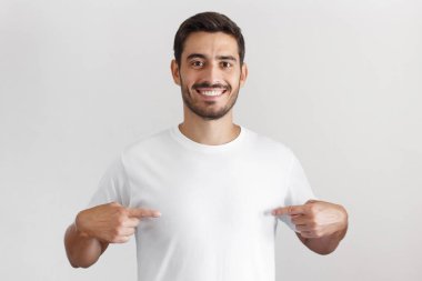 Boş beyaz t-shirt kopya alanı için reklam, her iki işaret parmakları ile işaret gülümseyen güzel adam gri arka plan üzerinde izole