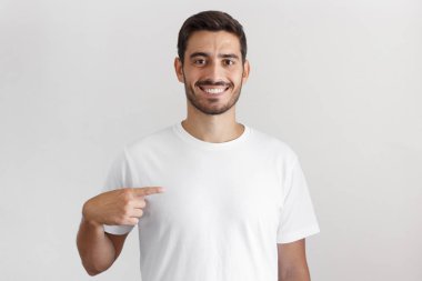 Gün ışığı atış onun boş beyaz t-shirt ile işaret parmağı, reklamlarınız için kopya alanı gösteren gülümseyen genç adamın gri arka plan üzerinde izole