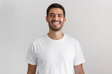 Doğruca kameraya mutlu gülümseme ile seyir, gülme boş beyaz casual t-shirt ayakta gri arka plan üzerinde izole olumlu Avrupa gencin yatay atış