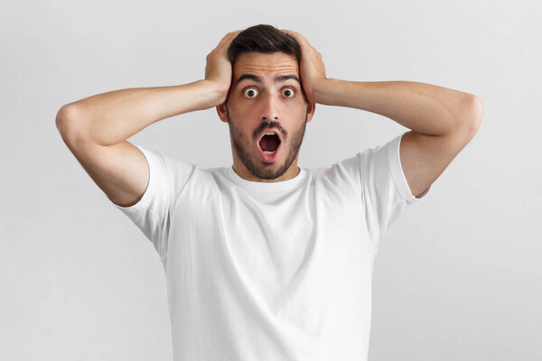 Daylight portrait of man isolated on gray background, with strong expression of fear, round eyes, open mouth and hands pressed to top of head, showing astonishment and shock