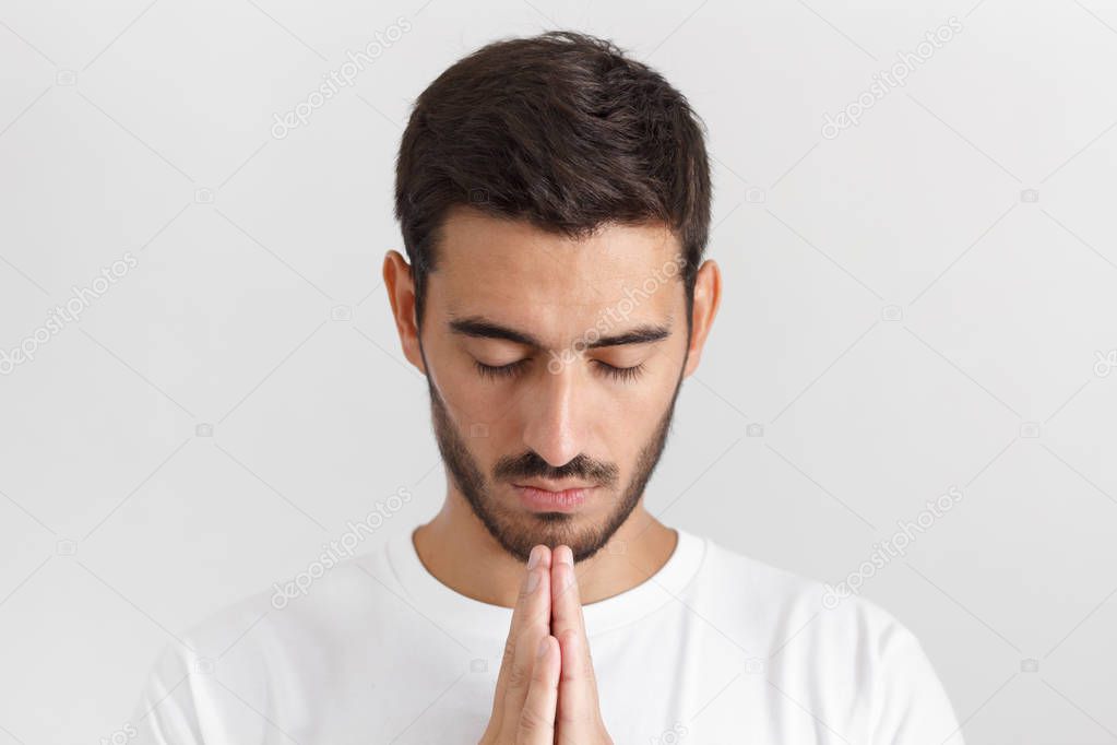 Daylight shot of young man in white tshirt isolated on gray background looking stressed, putting hands together as if he is praying with closed eyes 
