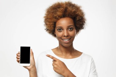 Boş smartphone gösteren ve ekrana işaret gri arka plan üzerinde izole beyaz t gömlek giymiş mutlu Afrikalı-Amerikalı kadın. Reklamcılık ürünlerimiz ve hizmetlerimiz için boşaltmak