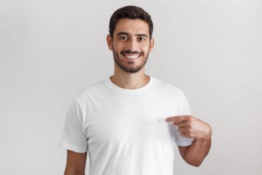 Beyaz boş t-shirt, boşaltmak için reklam işaret parmağı ile işaret ederken gülümseyen gri arka plan üzerinde izole genç yakışıklı beyaz adam yatay çekim