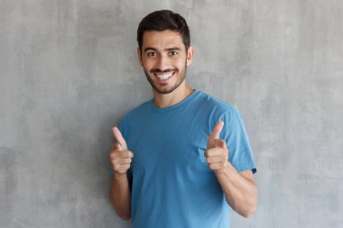 Genç adam açık ağız ile mutlu bir şekilde gülümseyerek ve kamera iki eliyle sanki onların çevirmek olduğunu söyleyerek işaret eden mavi t-shirt giyen gri dokulu duvar izole
