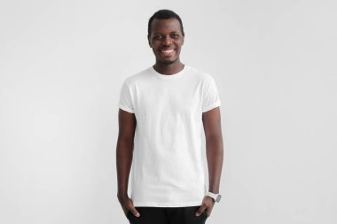 Afrika adam koyu tenli ayakta elleri ceplerinde ile gülümseyen Genç, logo veya metin, kopyalama yeri olan boş beyaz t gömlek giymiş izole gri arka plan üzerinde