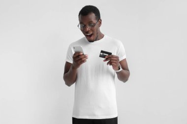 Genç yakışıklı Afrikalı-Amerikalı adam ayakta gri arka plan üzerinde izole smartphone bir el ve kredi kartı tutan, kablosuz bağlantı üzerinden ödeme işleme