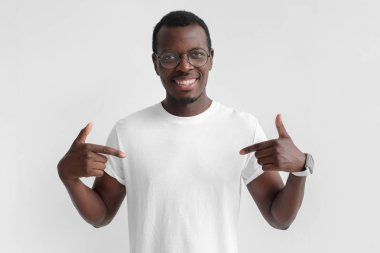 Boş beyaz t-shirt kopya alanı için reklam, her iki işaret parmakları ile işaret gülümseyen Afrikalı-Amerikalı adam gri arka plan üzerinde izole