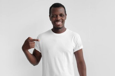 Onun boş beyaz t-shirt işaret parmağı ile işaret genç Afro-Amerikan adam gülümsüyor, alan gri arka plan üzerinde izole, reklamlarınız için kopyalama
