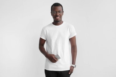 Açık gülümseme ile kameraya bakarak genç Afro-Amerikan adam beyaz t-shirt ve cep telefonu elinde tutan gri arka plan üzerinde izole gözlükler