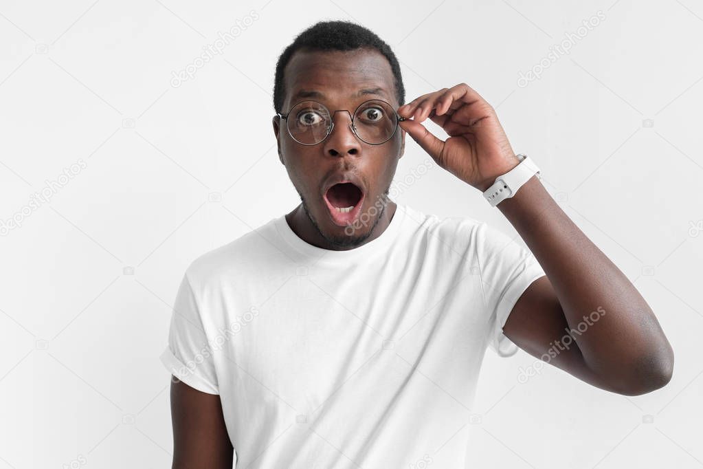 Closeup shot of young african american man wearing round eyeglasses, isolated on gray background, holding rim with fingers, looking really surprised and shocked with open mouth and round eyes