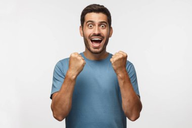 Zaferini kutluyor ve kazanan, mutluluk ve şans, derin duygusal ifade Yumruklarını sıkma sanki hareket gri arka plan üzerinde izole genç adam bağırarak