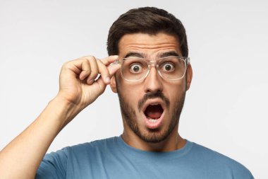 Oh Tanrım şeffaf gözlük, gri arka plan üzerinde izole tutarak açık ağız, düşük fiyat ve satış, tarafından sürpriz ile bağırarak mavi t-shirt genç adam