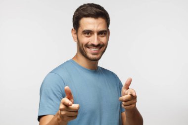 Hey siz! Mavi t-shirt kameraya gri arka plan üzerinde izole parmaklarıyla işaret eden genç adam