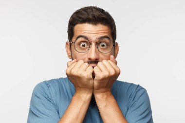 Young man isolated on gray background, covering mouth with hands and round eyes, wearing round eyeglasses, experiencing deep astonishment and fear clipart