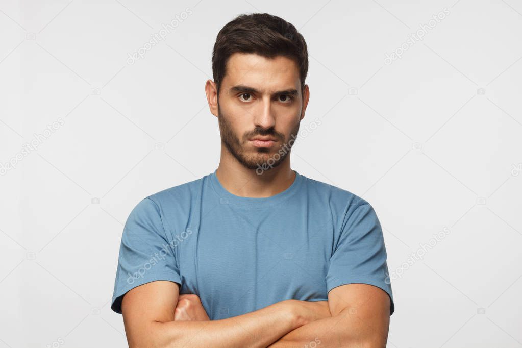 Young man in blue t-shirts tanding with arms crossed and serious concentrated face at camera, looking aggressive, isolated on gray background 