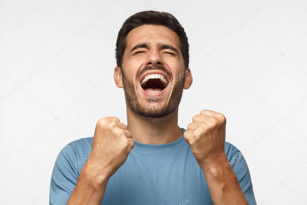 Young man isolated on gray background shouting with closed eyes, celebrating victory, squeezing fists in deep emotional expression of happiness and luck