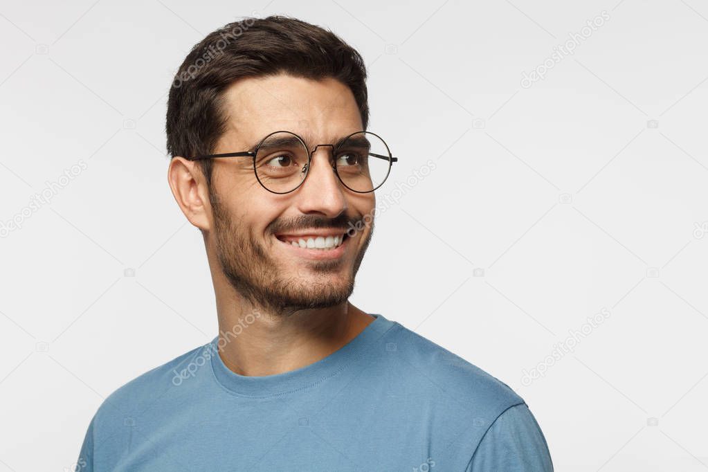 Closeup headshot of young man in round eyeglasses isolated on gray background, smiling happily, looking right, feeling positive, relaxed and joyful