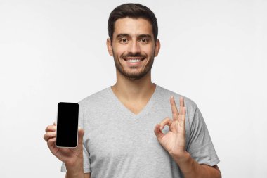 Genç adam boş smartphone ile boş siyah sreen tutarak ok ya da iyi jest gri arka plan üzerinde izole bir eliyle gösterilen gülümseyen