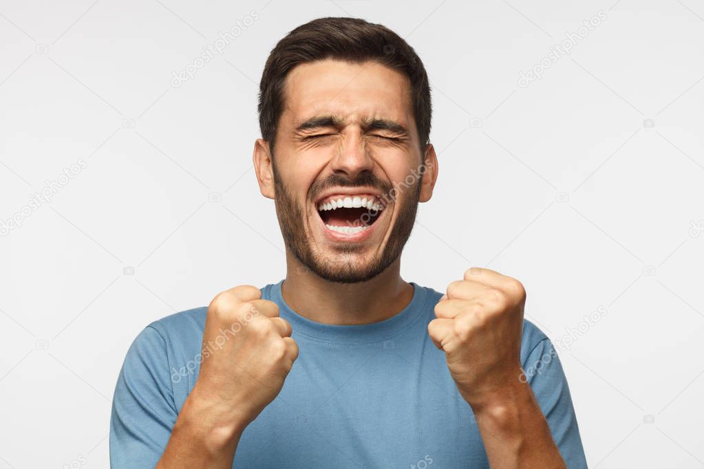 Closeup of emotional man isolated on gray background, screaming with joy and victorious expression, holding hands in gesture of winner, looking happy