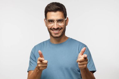 Hey siz! Mavi t-shirt ve şeffaf gözlük kamera için gri arka plan üzerinde izole parmaklarıyla işaret eden genç adam