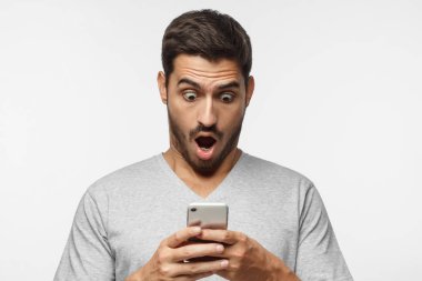 Şok içerik. Sergilenen bu onun akıllı telefon, web medya tarafından etkilendim arayan genç adam gri arka plan üzerinde izole portresi, heyecanlı. 
