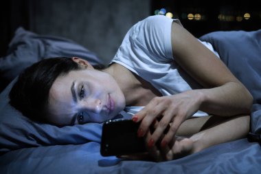 Akıllı telefon bağımlılığı. Genç, yorgun kadın cep telefonu ekranına bakıyor, gece geç saatte yatakta uzanıyor, sosyal medya haberlerine bakıyor.