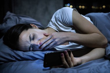 Açık ağız wih el kapanış cep telefonuyla yatakta, Genç esneme yorgun kadın