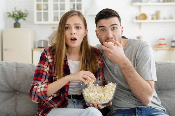 惊讶的年轻夫妇看电视坐在沙发上在家里与爆米花碗 — 图库照片