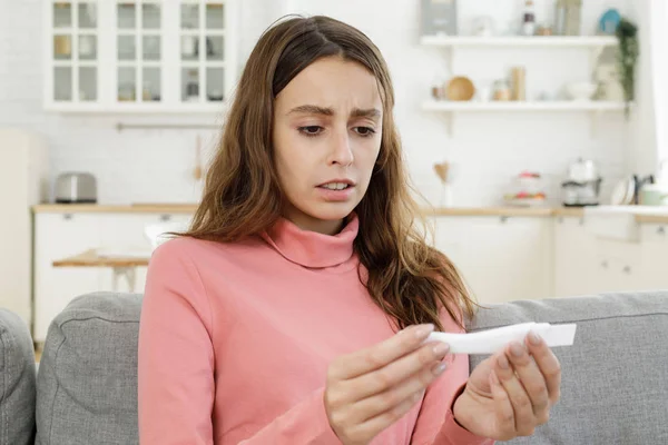 Unwanted pregnancy concept. Sad pregnant teen sitting on couch at home, checking a pregnancy test with positive result