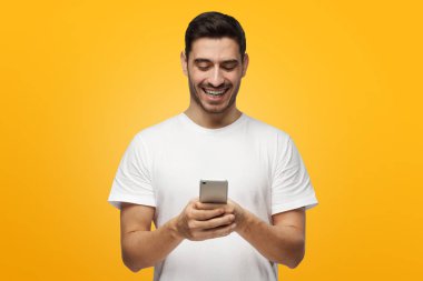 Yakışıklı beyaz adam gülerek mutlu bir şekilde her iki elinde tutan Smartphone ekranında içeriğe göre eğlendirdi beyaz casual T-shirt giyen gri arka plan üzerinde izole kapalı resmi